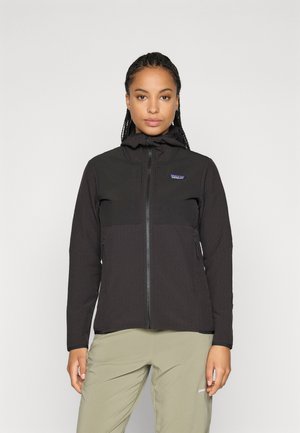 Kvinde med fletninger iført en sort zip-up jakke med hætte og lysegrønne bukser, stående foran en ensfarvet baggrund.