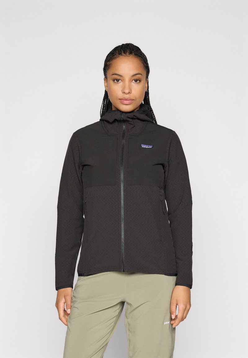 Woman with braided hair wearing a black zip-up jacket with hood and light green pants, standing against a plain background.