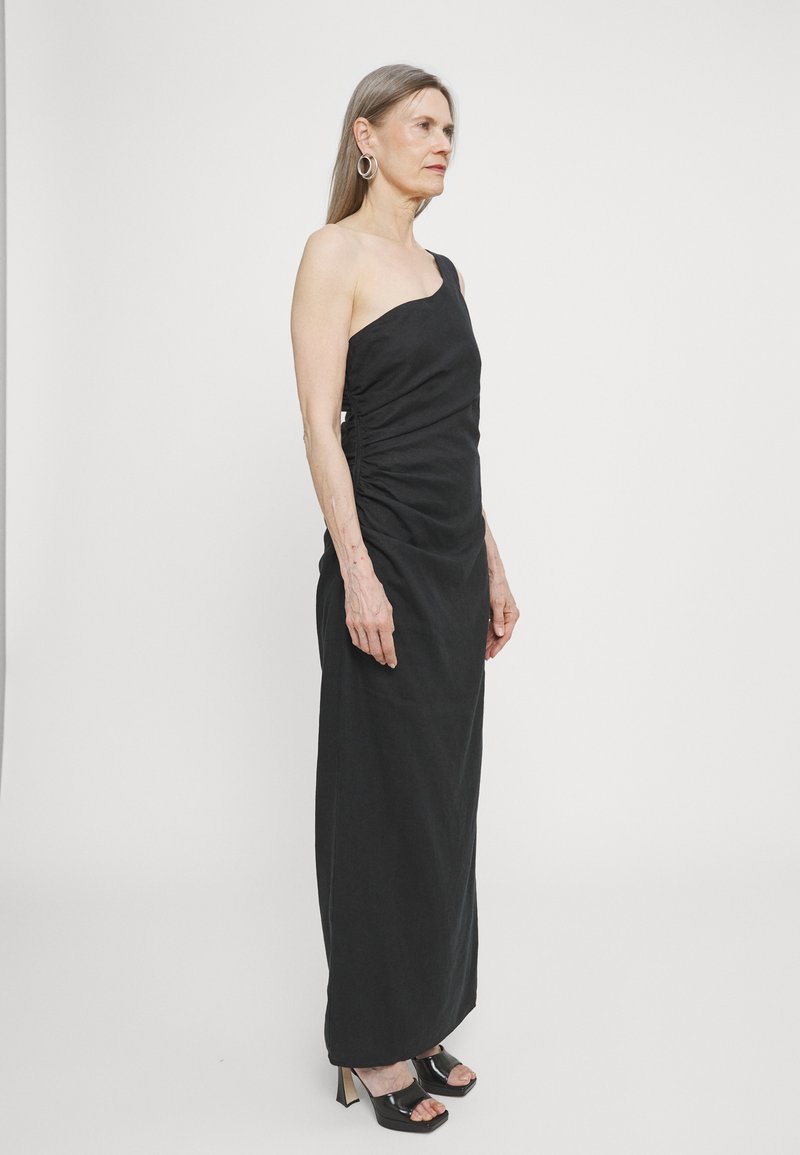 Black, one-shoulder dress with gathered side detailing, floor-length design, and smooth texture. Worn with black platform heels.