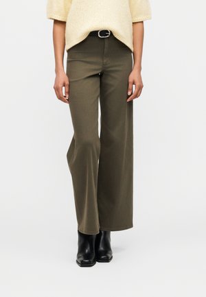 Personne portant un pantalon ample vert olive, une ceinture noire, des bottines noires et un pull en maille à manches courtes jaune.
