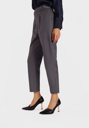 Pantaloni grigi su misura con vita alta e gamba dritta, abbinati a décolleté nere con punta appuntita e un accento dorato.