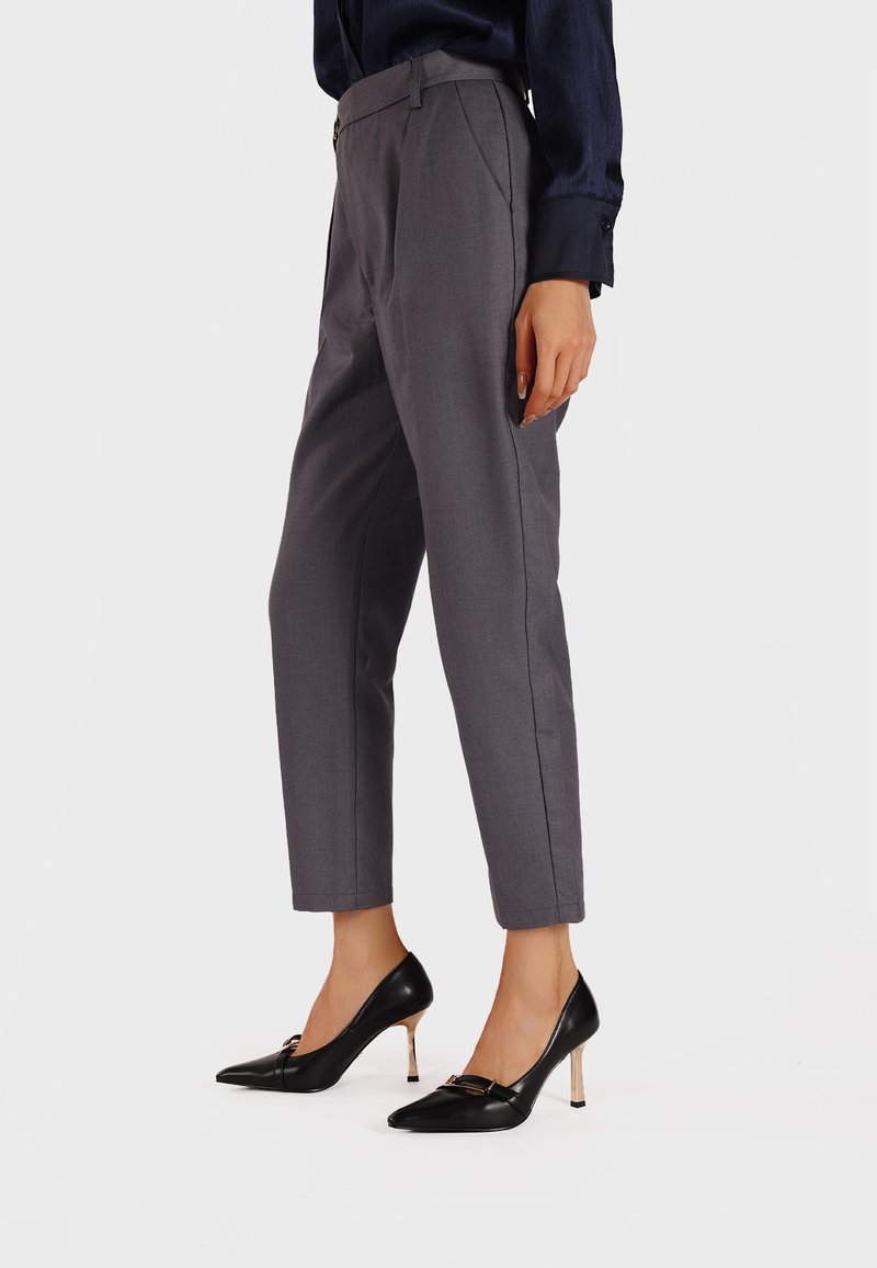 Pantaloni grigi su misura con vita alta e gamba dritta, abbinati a décolleté nere con punta appuntita e un accento dorato.
