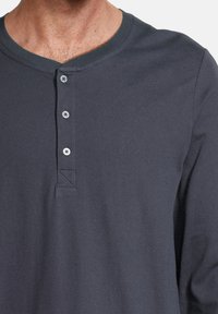 Chemise henley à manches longues gris foncé avec un col arrondi, quatre boutons et une texture lisse. Présente un détail de couture subtil sur la patte de boutonnage.