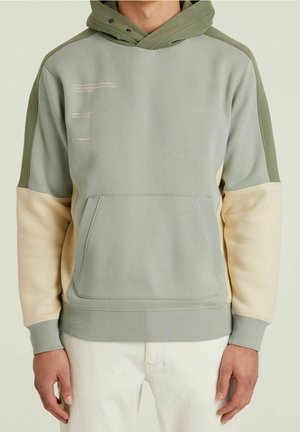 Mies, joka pukeutunut vihreän ja beigen sävyiseen kolmiväriseen hupulliseen huppariin, jossa on etutasku ja tekstitulostus vasemmassa ylärinnassa.