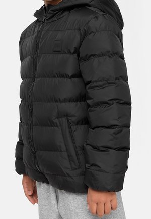 Sort quiltet hættejakke med frontlynlås og sidelommer båret af en person i grå bukser mod hvid baggrund.