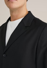 Musthave musta bleiser, millel on sile tekstuur, sooned kraed, ühe nööbiga sulgemine ja mis on kantud valge särke peal. Puhtad, struktureeritud jooned.