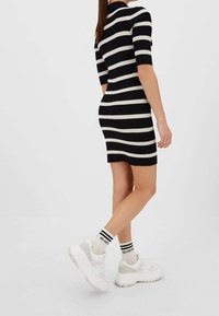 Robe en tricot rayée noir et crème, avec des manches courtes et une silhouette ajustée ; associée à des baskets blanches épaisses et des chaussettes rayées.