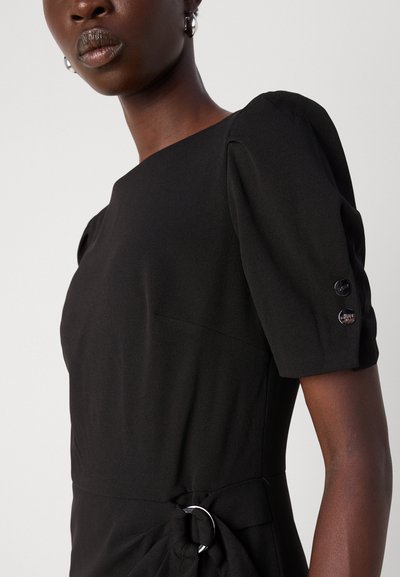 Robe noire avec des manches courtes bouffantes, un détail de nœud sur le côté, un accent en métal circulaire et une texture subtile. Elle présente un décolleté carré et des coutures minimales.