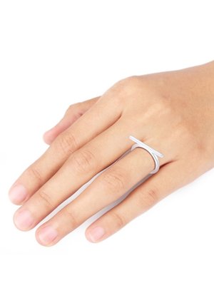 Main avec des doigts fins portant une bague argentée minimaliste avec une barre horizontale sur le majeur, sur fond blanc.