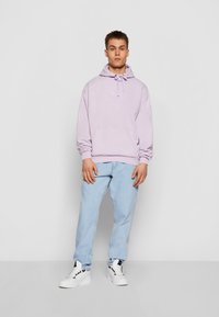 Felpa viola chiaro con tasca frontale, cordino e logo ricamato, abbinata a jeans azzurri e sneakers bianche.