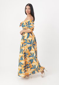 Schulterfreies Maxikleid mit Blumendesign in Gelb, Blau und Weiß, mit gerafften Ärmel und taillierter Taille, kombiniert mit Absätzen.