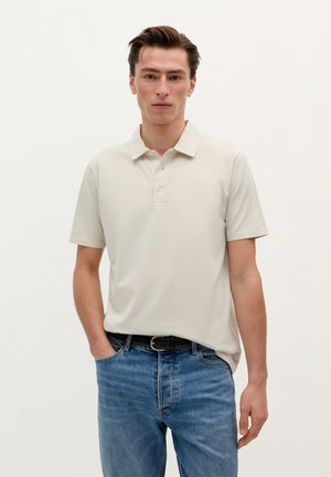 Polo shirt beige clair avec un col classique et trois boutons, associé à un jean en denim bleu. Il présente des manches courtes et une coupe décontractée.