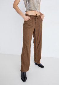 Bruna vida byxor med dragsko i midjan, som har sidofickor och synliga sömmar, tillsammans med svarta spetsiga skor.