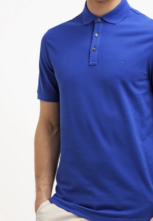 Mann trägt ein kurzärmeliges königsblaues Poloshirt mit drei Knöpfen und beige Hosen, steht mit einer Hand in der Tasche.
