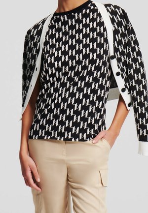 Person wearing a black and white patterned shirt and matching cardigan draped over shoulders, paired with beige pants with side pockets.