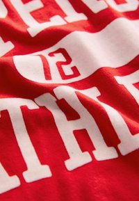 Rød bomuldsstof med en krøllet tekstur, med iøjnefaldende hvid skrift der inkluderer "12" og "TH." Designet fremhæver en sporty æstetik.