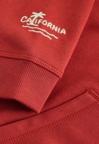Red sweatshirt fabric featuring a ribbed cuff and hem, with a white embroidered "CALIFORNIA" logo and palm tree design.