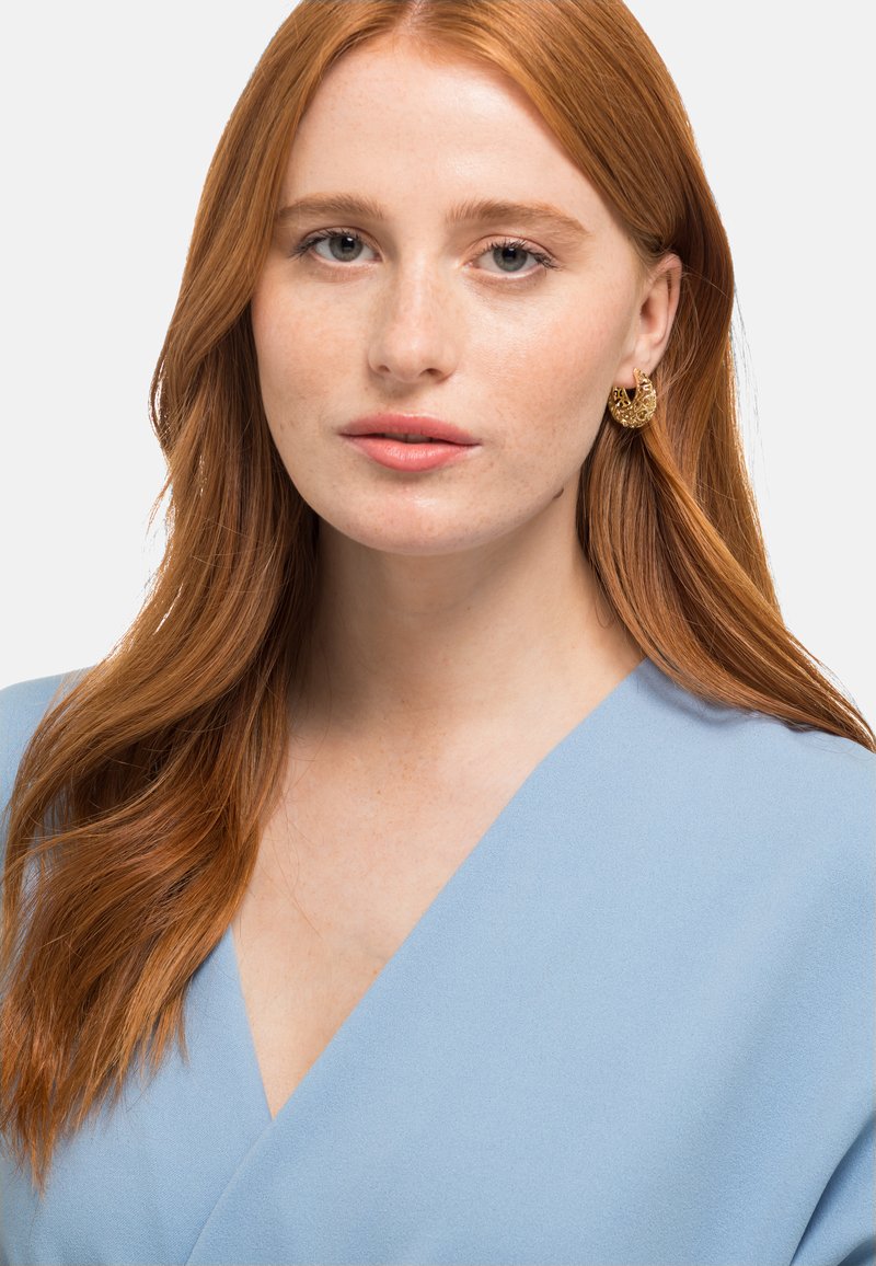 Top wrap bleu clair avec une texture douce, présentant un décolleté plongeant. Boucles d'oreilles dorées en forme de croissant avec un motif décoratif. Cheveux roux naturels.
