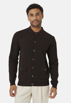Uomo che indossa un cardigan marrone scuro con trama, abbottonato, con tasche a patta e pantaloni beige, in piedi davanti a uno sfondo chiaro e uniforme.