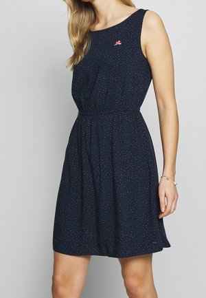 Kobieta w granatowej, bezrękawnikowej sukience w drobne białe kropki, z małą kolorową haftowaną kwiatową ozdobą na piersi.