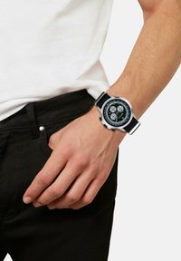 Montre chronographe noire avec un cadran rond, une lunette grise et des accents verts, accompagnée d'un bracelet en tissu. Portée sur une main contre une chemise blanche.