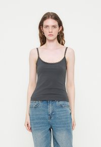 Jeune femme aux cheveux ondulés portant un caraco noir bordé de dentelle et un jean bleu clair, debout devant un fond blanc uni.