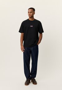 Άνδρας στέκεται με το χέρι στην τσέπη φορώντας μαύρο γραφικό μπλουζάκι, σκούρο μπλε τζιν και καφέ δερμάτινα παπούτσια πάνω σε κρεμ φόντο.