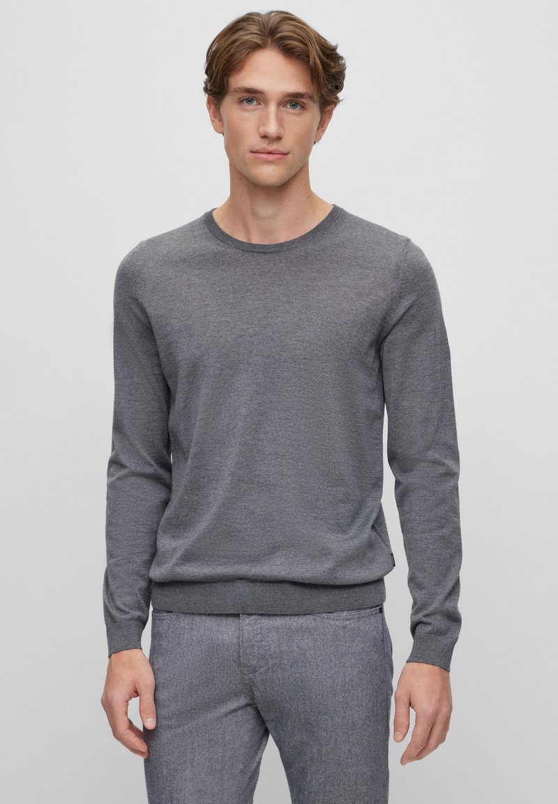 Grauer Langarmpullover mit rundem Hals, weichem Material und geripptem Saum. Kombiniert mit hellgrauen, maßgeschneiderten Hosen.