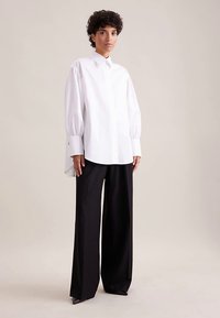 Camicia bianca a maniche lunghe con collo strutturato, caratterizzata da maniche voluminose e orlo asimmetrico. Abbinata a pantaloni neri a gamba larga.