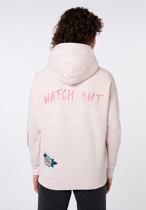 Person mit lockigem Haar trägt einen hellrosa Hoodie mit hochgezogener Kapuze, Text „PASS AUF, ERSTAUNLICHE DINGE KOMMEN“ und einen Blumenaufnäher auf dem Rücken.