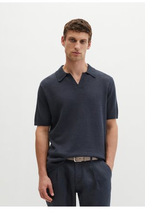 Mann trägt dunkelmarineblaues kurzärmeliges Strick-Poloshirt, leicht in marineblaue Hose mit beigem Gürtel gesteckt, steht vor einem einfarbigen weißen Hintergrund.