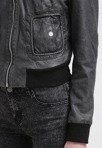 Giubbotto in pelle nera con zip e tasca quadrata dotata di bottone a pressione in metallo, indossato sopra dei jeans neri lavati in acido.