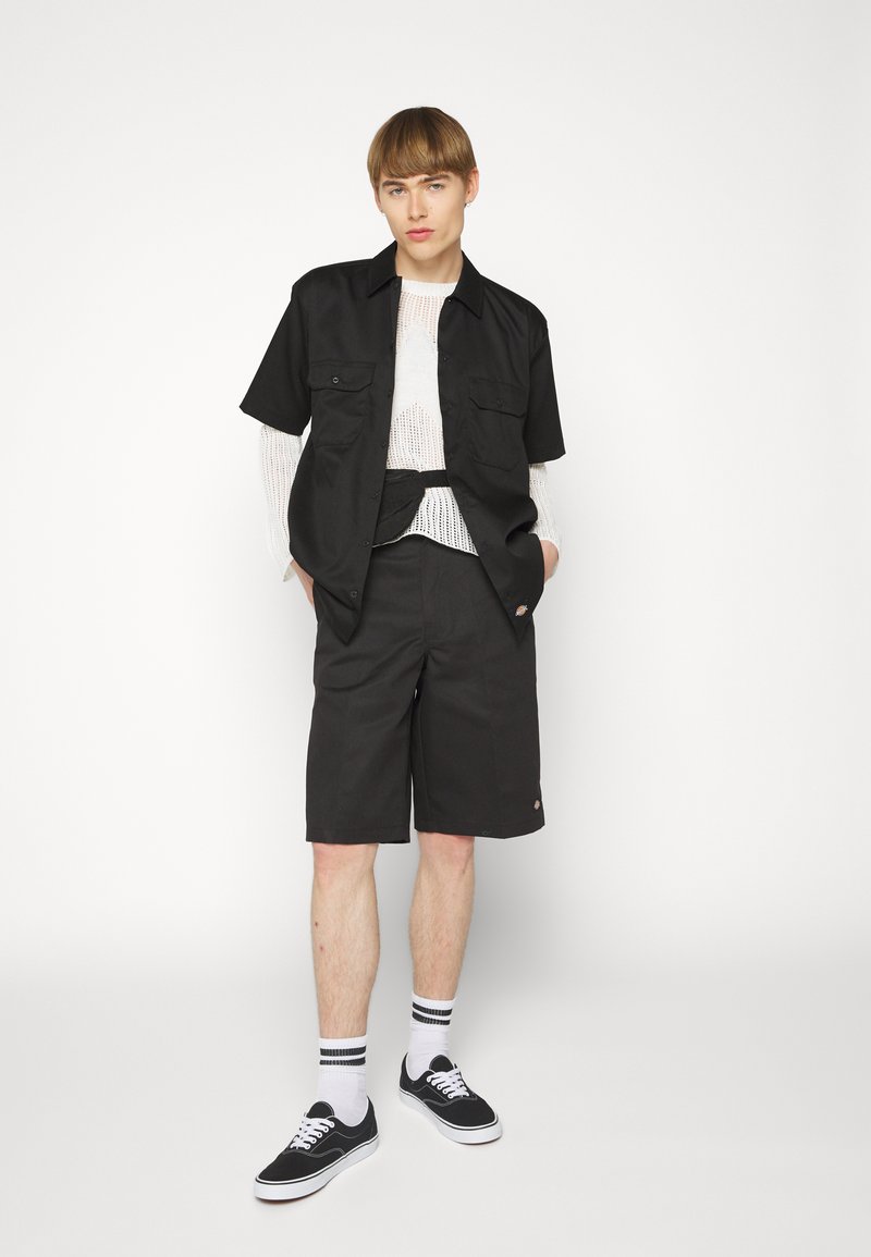 Black short-sleeve button-up shirt with two chest pockets, paired with black shorts. Model wears black sneakers with white laces and striped socks.