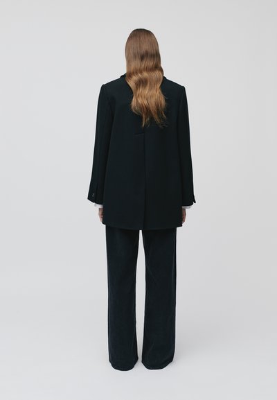Blazer oversize nero su misura con spalline, e pantaloni larghi scuri, entrambi con una texture liscia. I capelli cadono in morbide onde.
