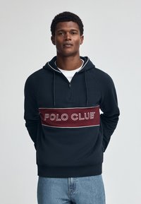 Felpa con cappuccio blu navy con zip a quarto, caratterizzata da una striscia orizzontale bordeaux con "POLO CLUB" in lettere bianche. Vestibilità casual, texture morbida.