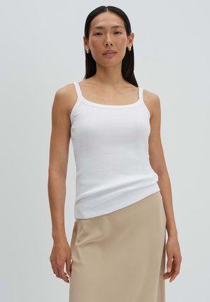 Femme aux cheveux longs et foncés portant des boucles d'oreilles argentées en anneau, un débardeur blanc côtelé et une jupe beige, sur un fond clair uni.