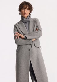 Kobieta z krótkimi brązowymi włosami, ubrana w długi szary płaszcz i szary sweter z golfem, stoi z założonymi rękami na tle gładkiego białego tła.