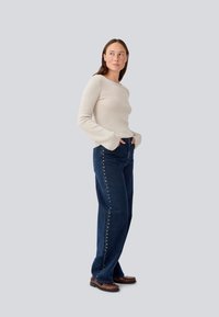 Beige ribbed long-sleeve top paired with high-waisted dark blue jeans featuring metallic button detailing along the side.