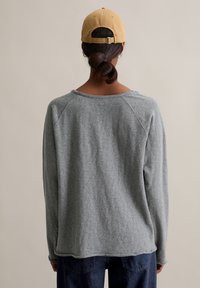 Szary d�ługi rękaw sweter z luźnym krojem, raglanowymi rękawami i surowym wykończeniem, zestawiony z beżową czapką bejsbolową z tyłu.