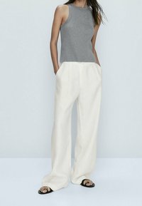 Top sans manches gris à côtes, col haut, associé à un pantalon en lin blanc ample avec poches, porté avec des sandales noires à glissière.