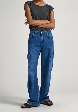Osoba stojąca ze skrzyżowanymi ramionami, ubrana w czarną bezrękawnik, niebieskie spodnie cargo i czarne sandały z odkrytymi palcami na jednolitym tle.