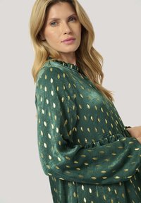 Grünes Kleid mit einem Rüschenausschnitt, langen Ärmeln und goldenen Blattmotiven. Weicher Stoff mit einem gerafften Taillendesign.