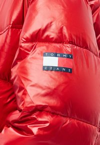 Κόκκινο μπουφάν τύπου puffer με γυαλιστερή, λεία υφή. Περιλαμβάνει ορθογώνιο λογότυπο της μάρκας με τις λέξεις "TOMMY" και "JEANS" σε αντιθετικά χρώματα.