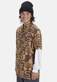 Korte mouw shirt in bruin en tan, met een gedurfde botanische print. Inclusief een borstzak en is gelaagd over een wit langemouwenshirt.