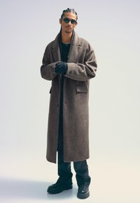 Hnedý vlnený kabát s voľným strihom, textúrovaným povrchom, nosený nad čiernym topom a nohavicami, doplnený čiernymi rukavicami a robustnými čiernymi čižmami.