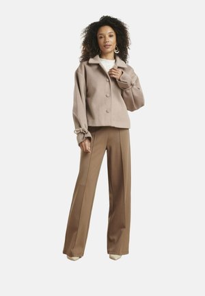 Vrouw met krullend haar, die een beige jasje met knopen en bruine wijde broek draagt, staand tegen een witte achtergrond.