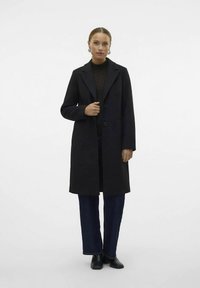 Sieviete ar garu melnu mēteli pār tumšas rakstainas blūzes, tumši ziliem džinsiem un melniem potītēm zābakiem, stāvot uz vienkārša fona.