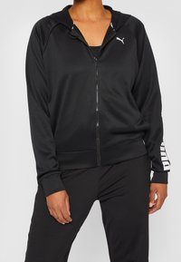 Sudadera con cremallera negra hecha de tela suave, con capucha, bolsillos laterales y acentos de logotipo blanco en el hombro y la manga.