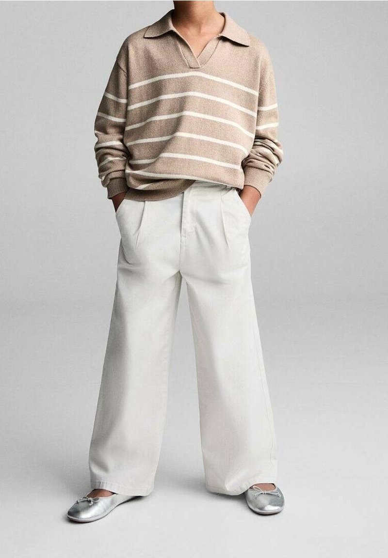 Personne portant un pull beige rayé à manches longues, un pantalon blanc à jambes larges et des ballerines argentées métalliques, debout avec les mains dans les poches.