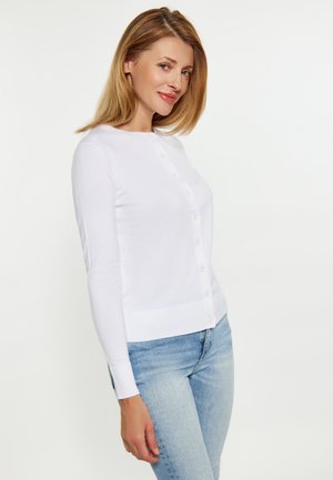 Kvinde med blondt hår iført en hvid knappet cardigan og lyseblå jeans, stående foran en ensfarvet hvid baggrund.